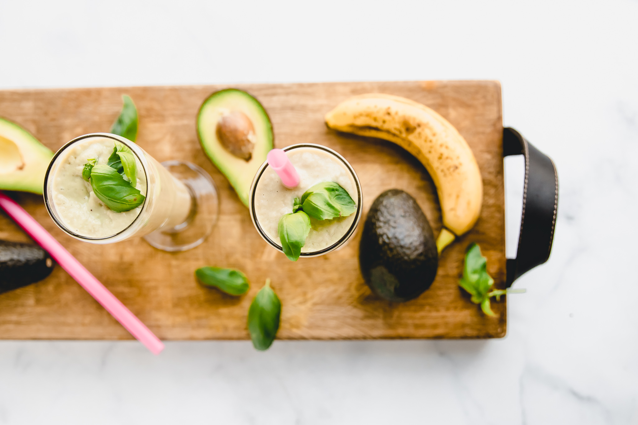 Avocado Basil Morning Smoothie. Made with avocado, basil, banana,pineapple, and coconut milk. Smoothies are on a William Sonoma charcuterie cutting board