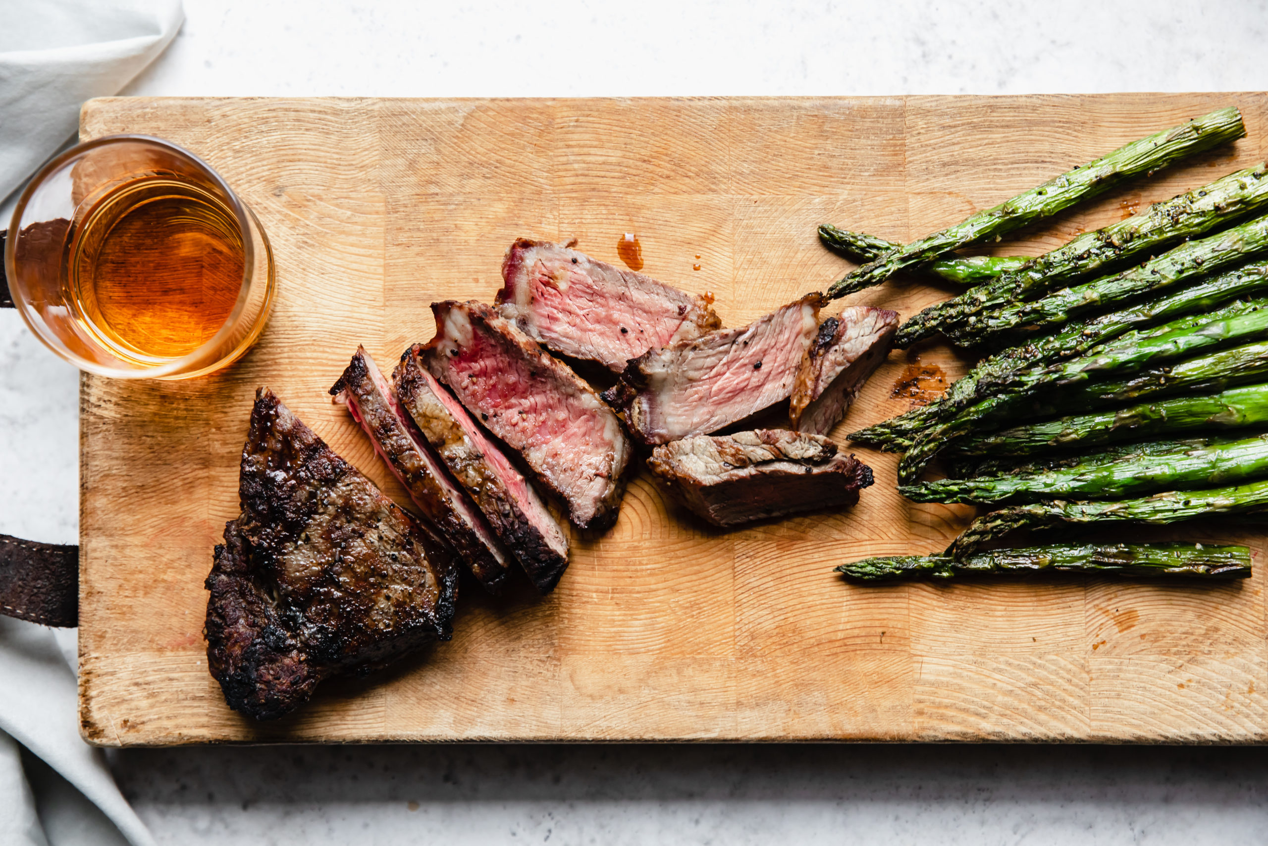 grilled steak cut into strips on a wooden bard with whiskey glass and asparagus on the side